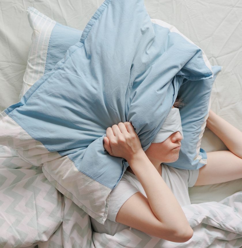woman with dark hair in bed with a mask over her eyes can't sleep and covers her head with pillows